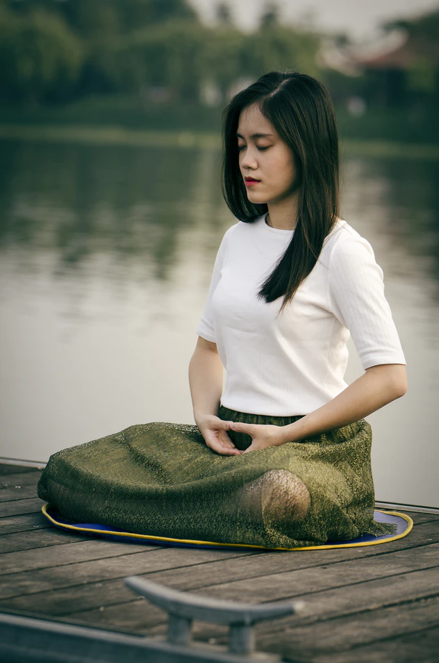Person meditating by a lake with eyes closed and wearing a white shirt and green bottoms.